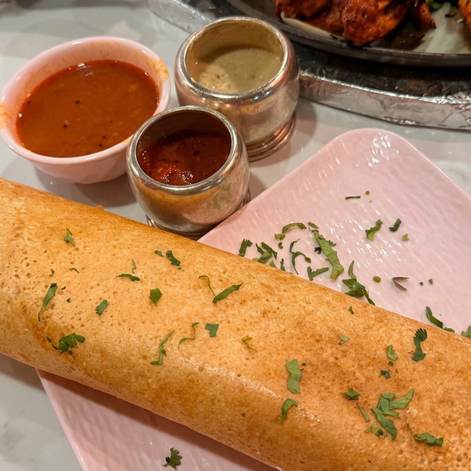Crisp golden dosa garnished with cilantro, served on a pink plate with three dipping sauces and grilled appetizers.