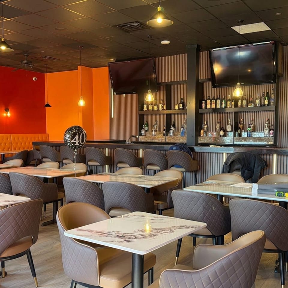 Modern restaurant dining room with orange walls, tufted banquette seating, marble tables, upholstered chairs, pendant lights, and stocked bar shelves.